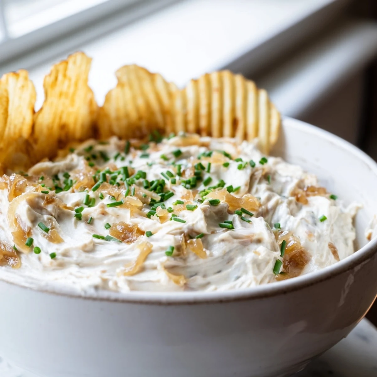 Creamy Game Day Onion Dip in a rustic bowl, with golden caramelized onions stirred into sour cream, served beside a pile of crispy potato chips.