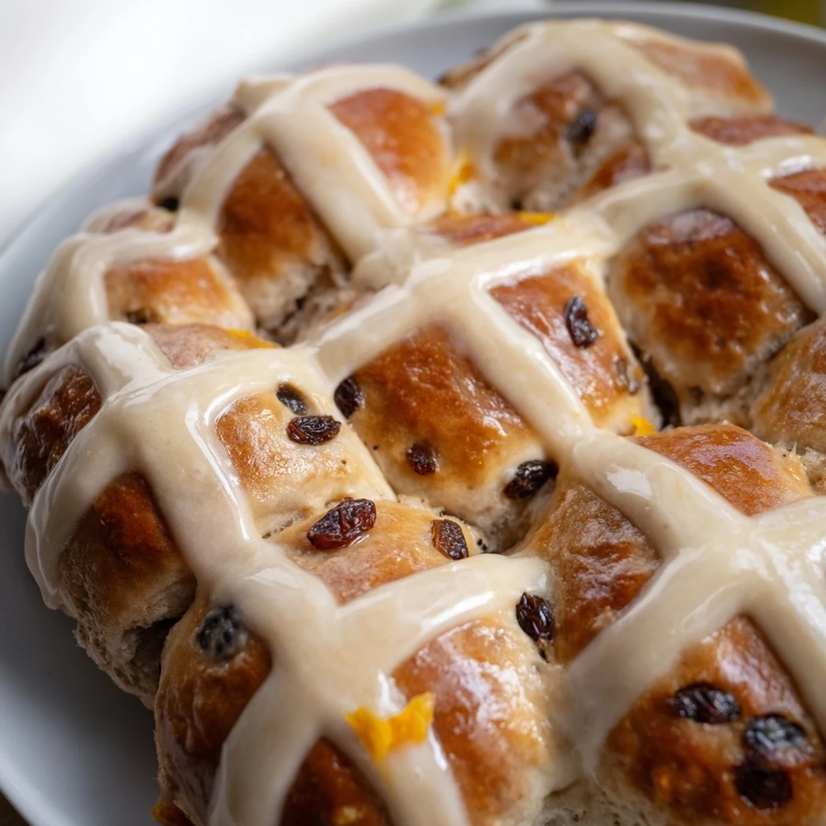 A close-up of Hot Cross Buns with Raisins and Orange Glaze showing the golden crust and glossy icing on a rustic wooden board.