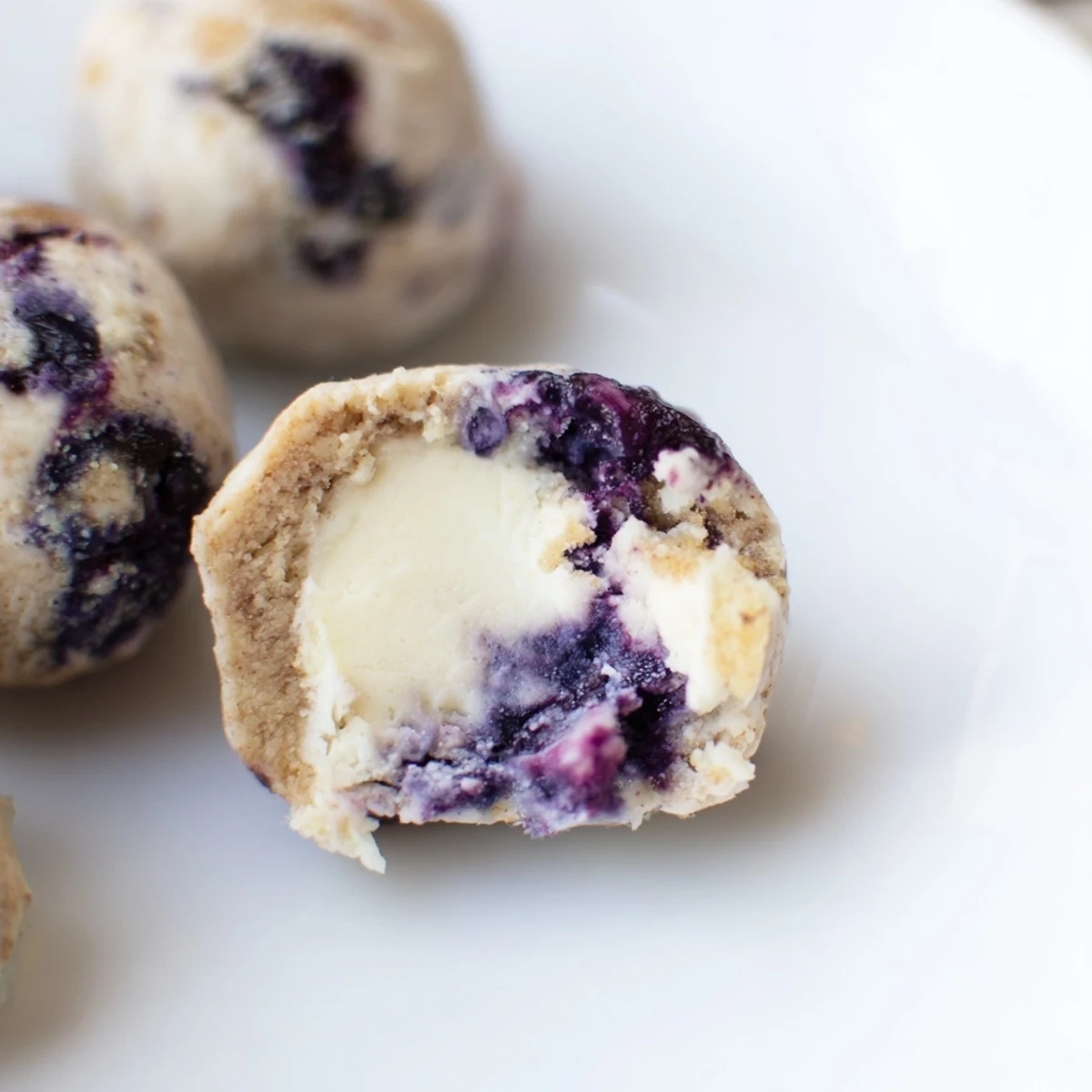 Freshly chilled Blueberry Cheesecake Protein Bites topped with a vibrant berry swirl rest on a white plate, showcasing their creamy texture.  