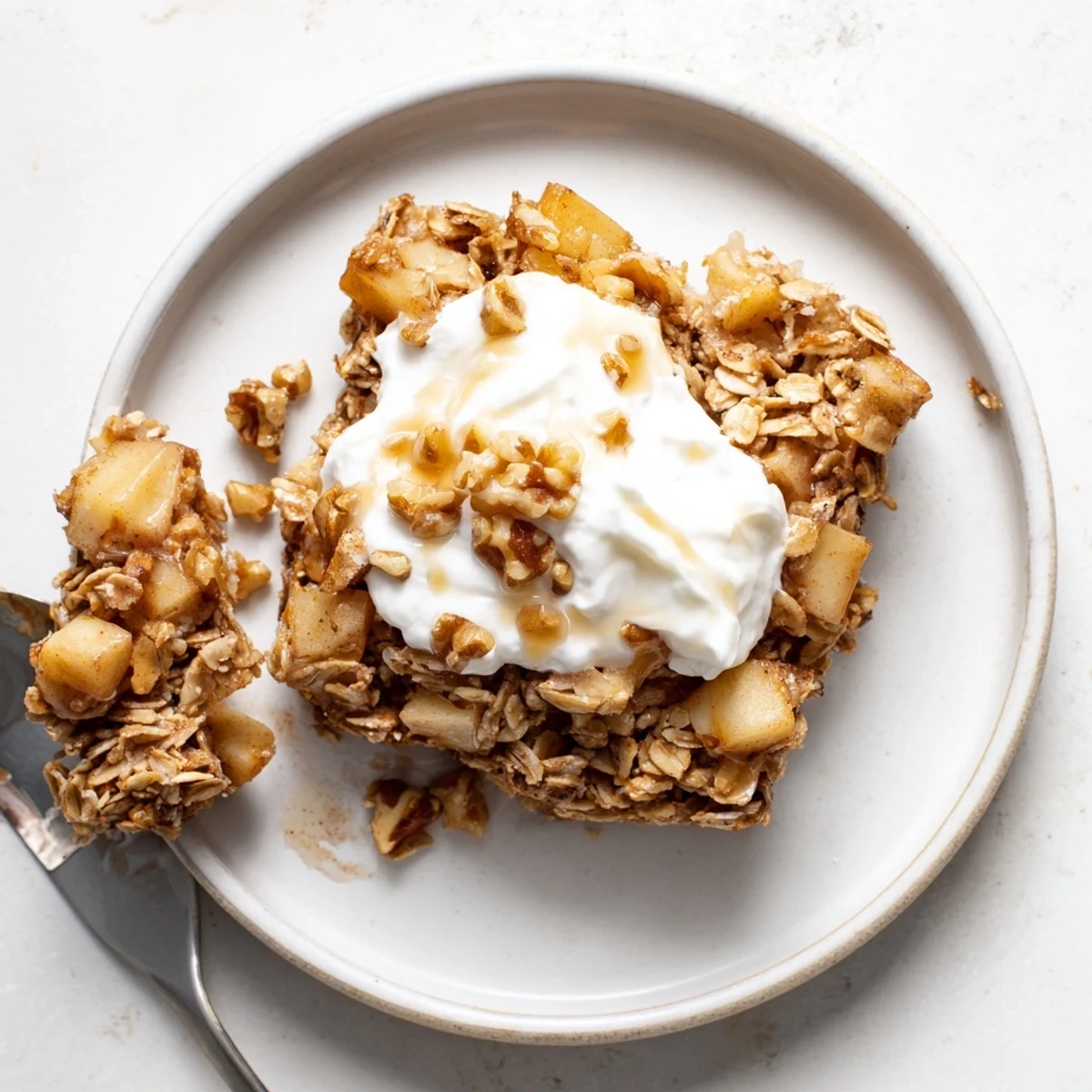 In a white dish, Baked Apple Oatmeal with Greek Yogurt is served warm, with a dollop of yogurt and pecans.