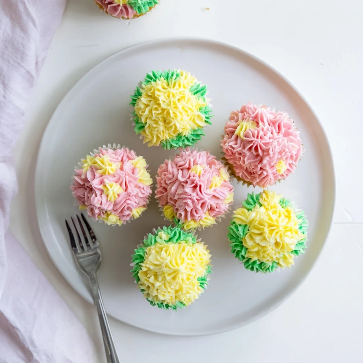 A close-up view of Baby In Bloom Cupcakes shows fluffy vanilla cake and swirls of floral buttercream, with soft pink and yellow petals and a hint of green leaves.