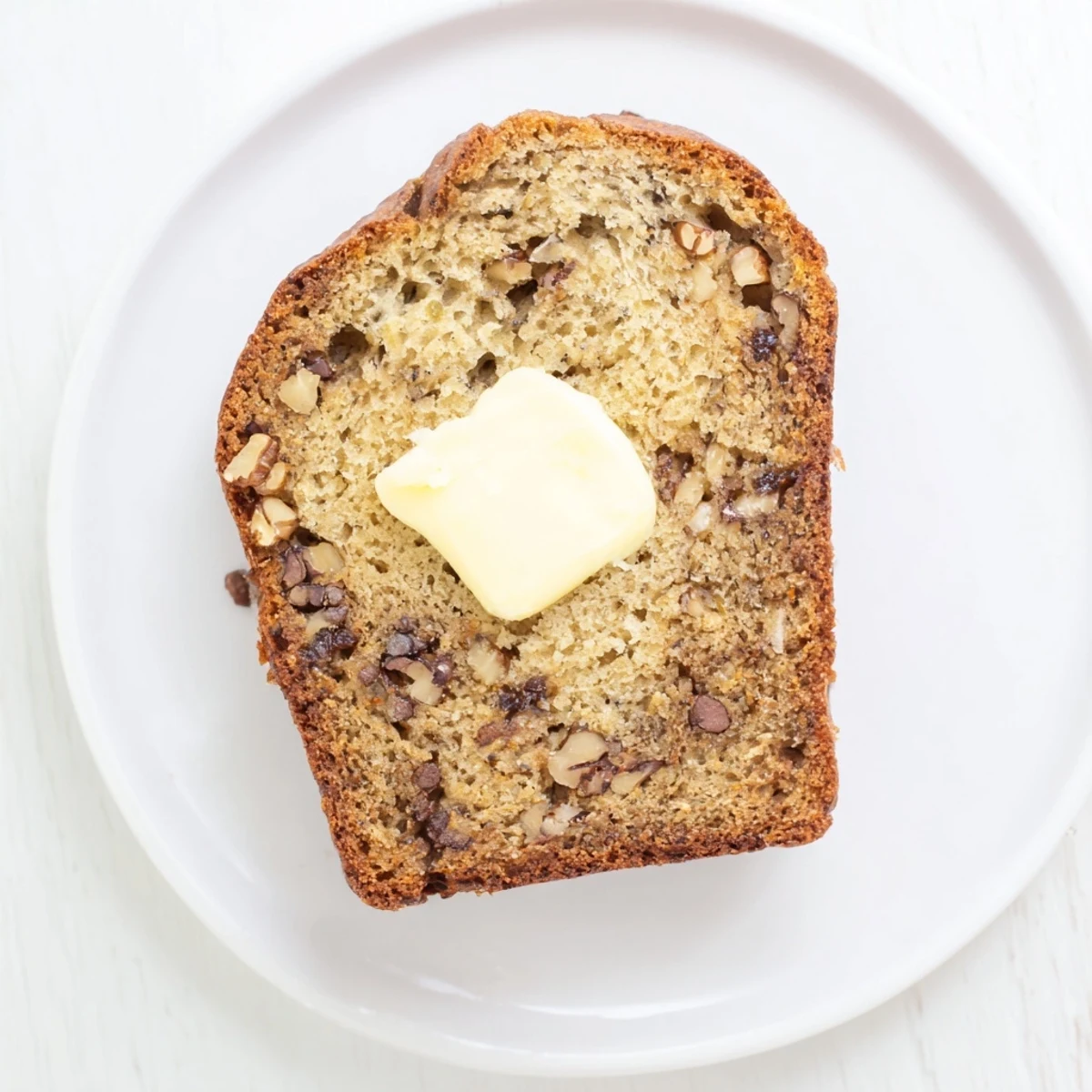 Thick slice of moist banana bread with visible banana flecks, served on a white plate with a pat of melted butter
